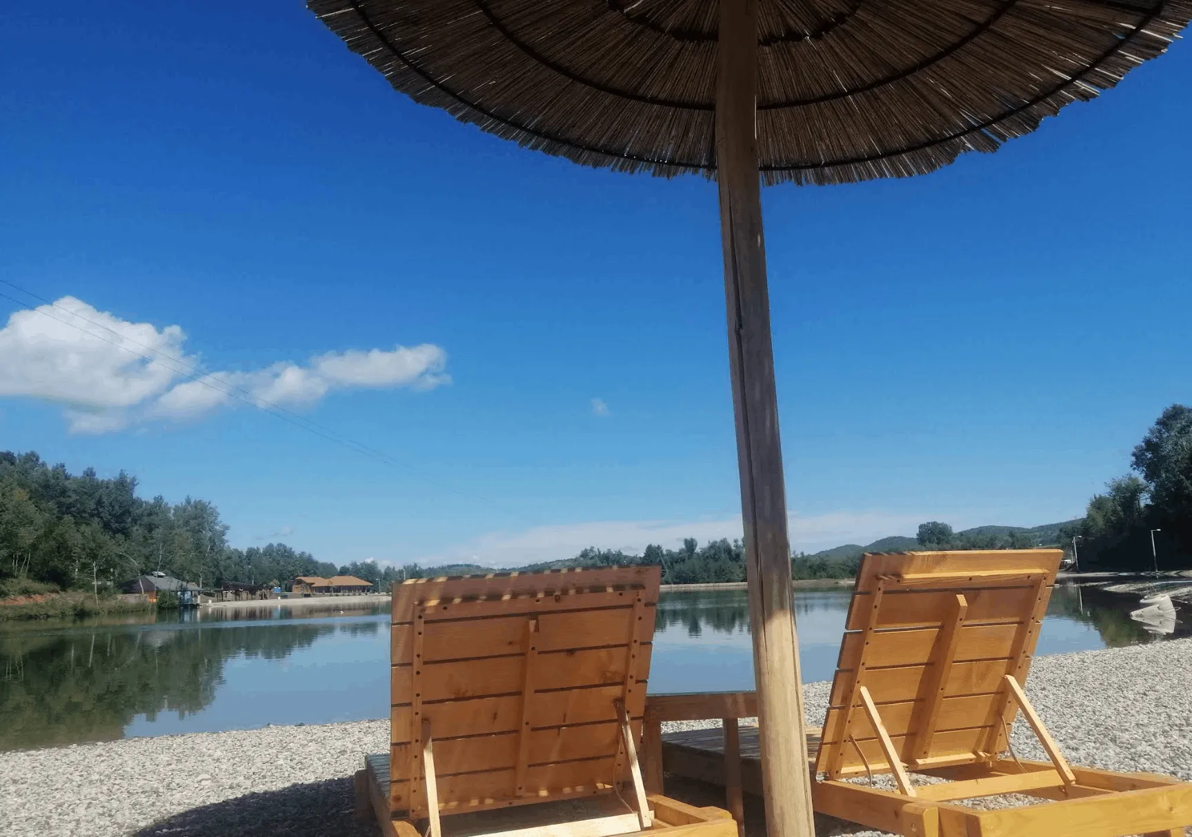 Wooden loungers by the lake on Manjača, with straw shade and a pebbled shore
