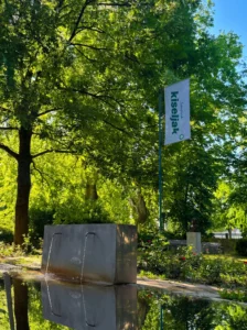 Düzenlenmiş bir parkta, açık akışlı doğal mineral suyu bulunan taş çeşme; etrafı sık yeşil ağaçlarla çevrili ve arka planda Sarajevski kiseljak markasının bayrağı yer alıyor