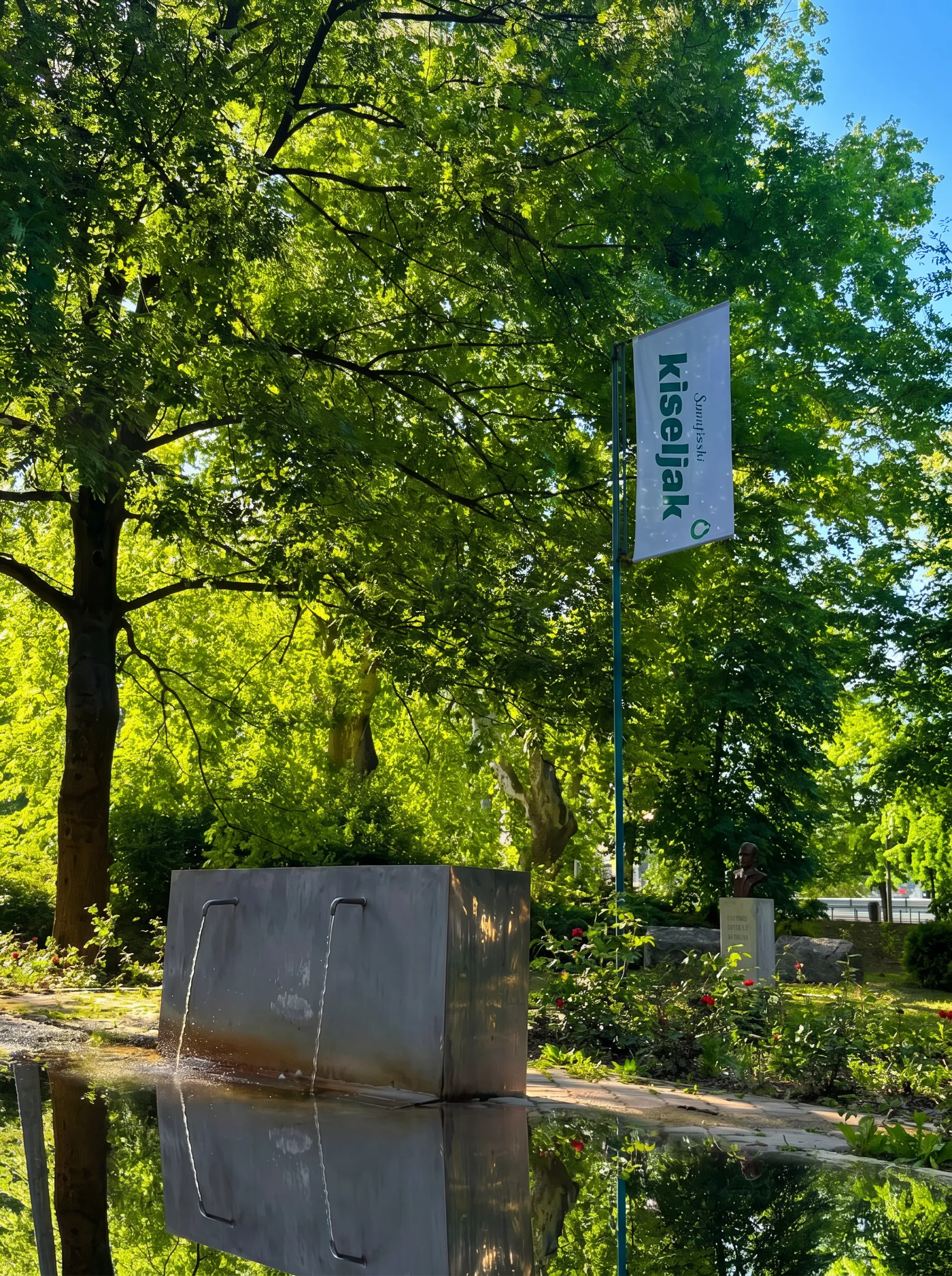 Düzenlenmiş bir parkta, açık akışlı doğal mineral suyu bulunan taş çeşme; etrafı sık yeşil ağaçlarla çevrili ve arka planda Sarajevski kiseljak markasının bayrağı yer alıyor
