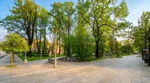 Pogled na široku popločanu pješačku zonu unutar parka okruženu bogatim zelenim grmljem, visokim drvećem i modernom uličnom rasvjetom