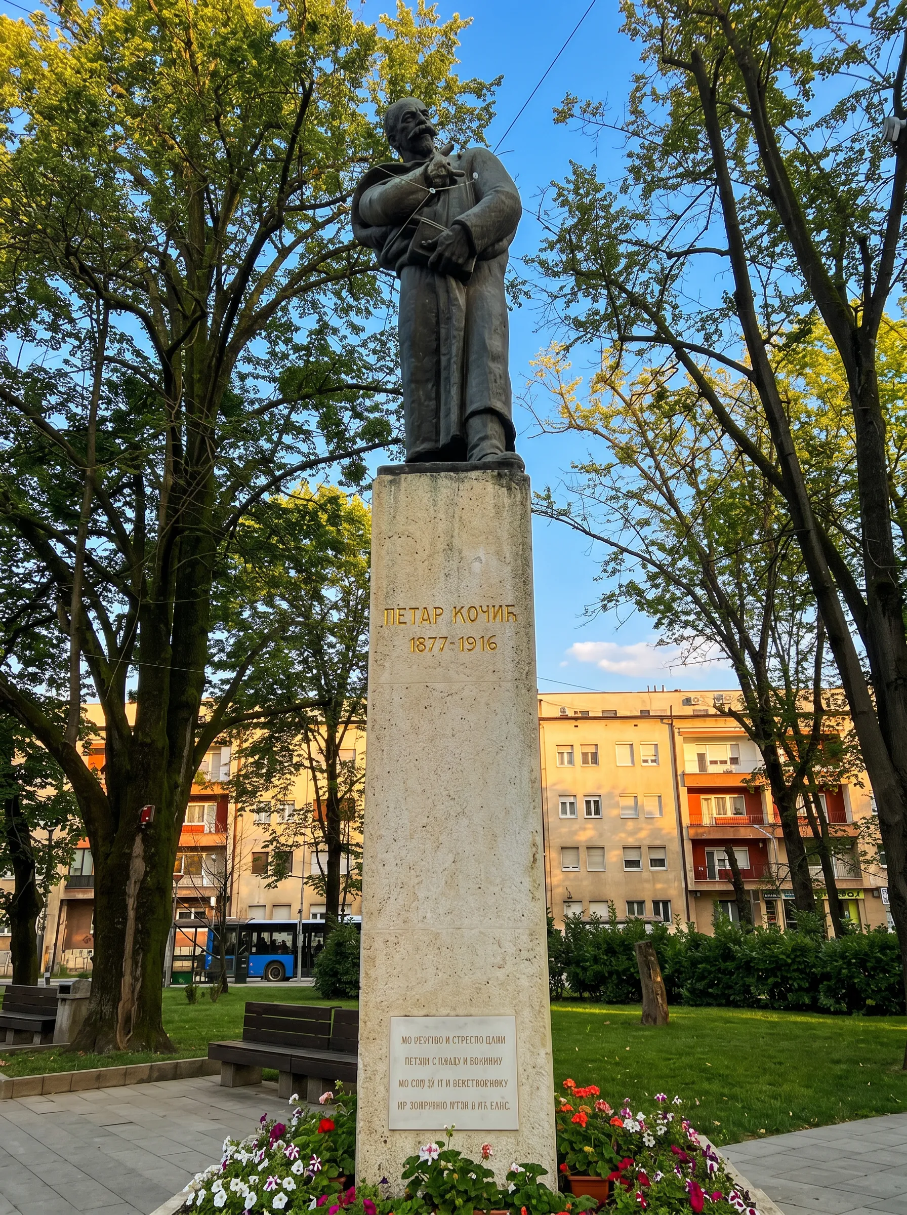 Visoki kameni postolje sa bronzanom statuom književnika Petra Kočića, smješteno u uređenom gradskom parku sa drvećem i zgradama u pozadini