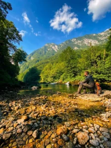 Un uomo siede sulla riva ghiaiosa e osserva la superficie calma del fiume Tara, in cui si riflettono gli alberi verde intenso e le alte catene montuose sullo sfondo