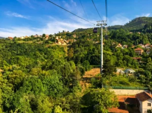 Sarajevo teleferiğinin kabini ve taşıyıcı direğinin, Trebević yamaçlarındaki sık yeşillik ve yerleşimlerin üzerinde yükselirken görünümü