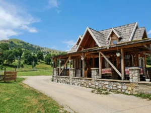 Tradicionalen gorski objekt, zgrajen iz lesa in kamna z urejeno teraso ob vaški cesti, obdan z zelenimi hribi pod modrim nebom