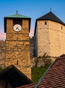 Detaillierter Blick auf den markanten steinernen Uhrturm mit Uhr und den massiven Turm der Festung in Tešanj unter einem Abendhimmel in zarten Rosatönen