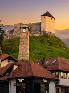 Historischer steinerner Uhrturm und die Mauern der Festung Tešanj auf einem grünen Hügel über den traditionellen roten Dächern der Häuser im alten Teil von Tešanj bei Sonnenuntergang