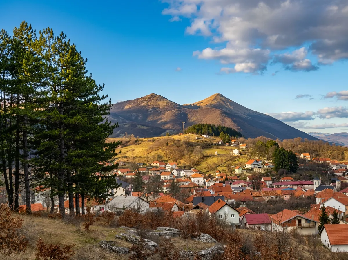 Pogled sa uzvišenja na crvene krovove kuća u naselju, sa prostranim zelenim i žutim brdima u pozadini iznad kojih se uzdižu planinski vrhovi pod plavim nebom i bijelim oblacima