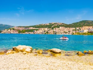 A person rides a jet ski, leaving a white trail across the turquoise-blue sea surface, photographed from the pebbly shore of Meredek Beach with a view of the town in the background