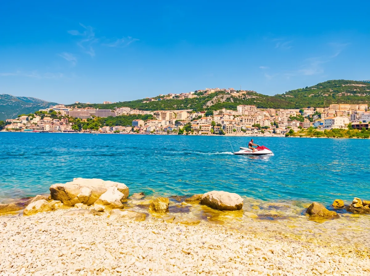 A person rides a jet ski, leaving a white trail across the turquoise-blue sea surface, photographed from the pebbly shore of Meredek Beach with a view of the town in the background