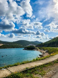 View from the hilltop and coastal path over the calm, semicircular cove of Meredek Beach, surrounded by green Mediterranean hills under a mostly cloudy sky